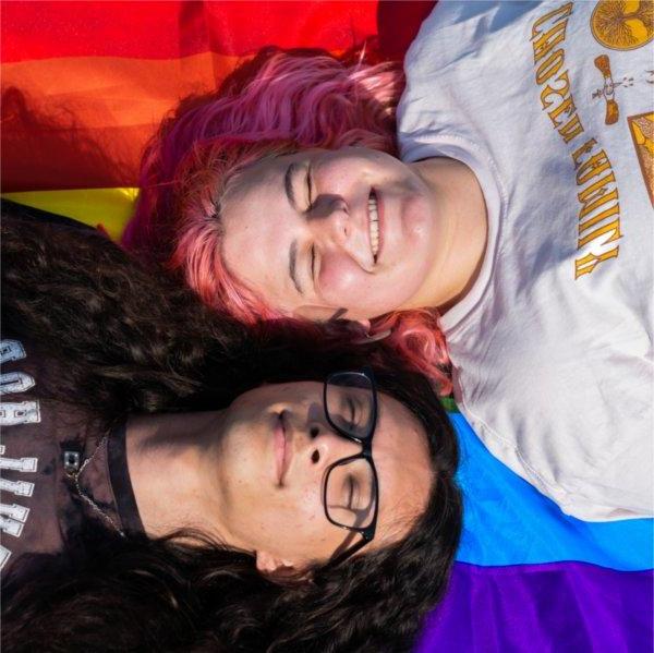 两个人，一个是粉色头发的, 另一个留着黑色长发，戴着眼镜，躺在彩虹骄傲旗上，对着镜头咧嘴笑着.