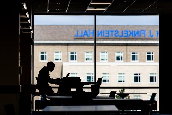  A student working on their laptop is silhouetted against a window in an academic building. 