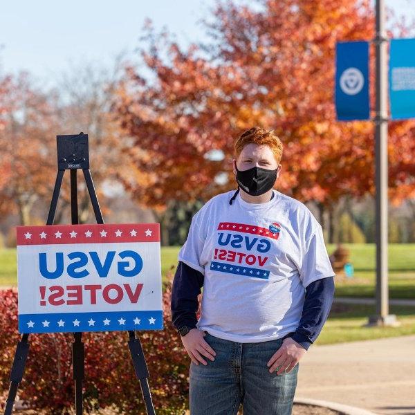 Sam Jacobs in mask standing outside next to GVSU Votes sign and wearing the same t-shirt, with GVSU Votes!