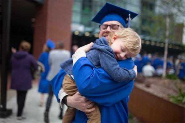  毕业典礼前，一位戴着学士帽、身穿学士服的毕业生抱着蹒跚学步的儿子.